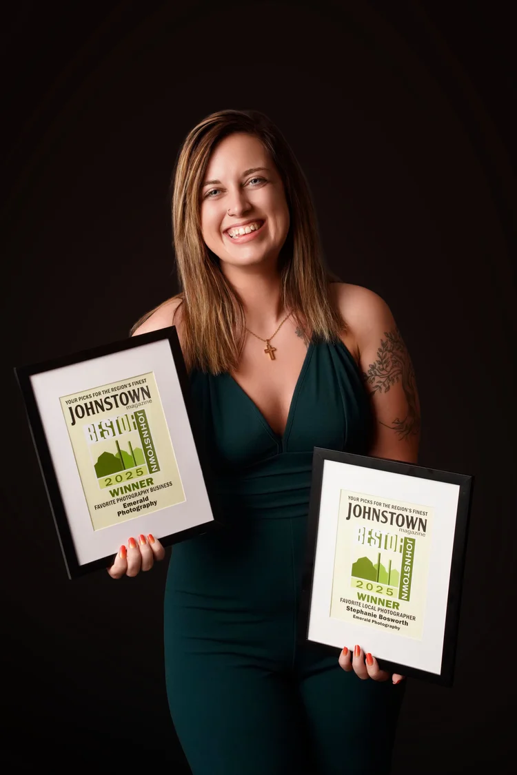 Stephanie Bosworth smiling in a dark green jumpsuit and holding two framed Best of Johnstown 2025 winner awards: Favorite Photography Business for Emerald Photography and Favorite Local Photographer