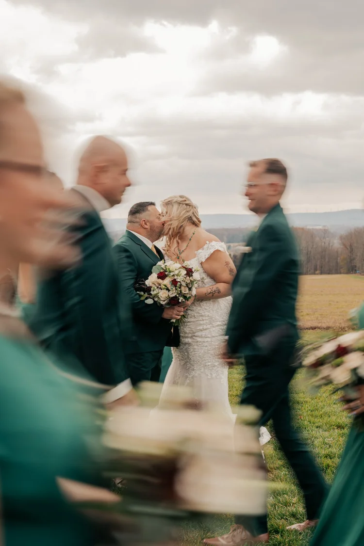 A bride and groom share a kiss outdoors during their wedding, surrounded by blurred wedding guests, with overcast skies and a grassy field in the background.