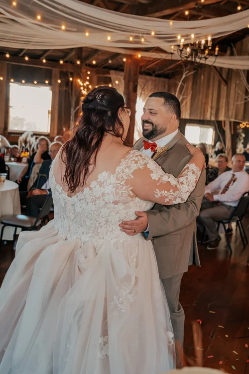 A bride and groom dancing at their wedding reception inside a rustic barn with string lights, surrounded by seated guests.